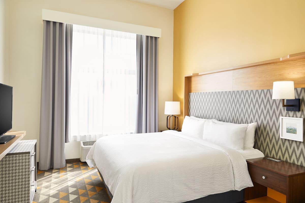 A hotel room with a large bed, a desk with a flat-screen TV, a bedside table with lamps, and a patterned floor.