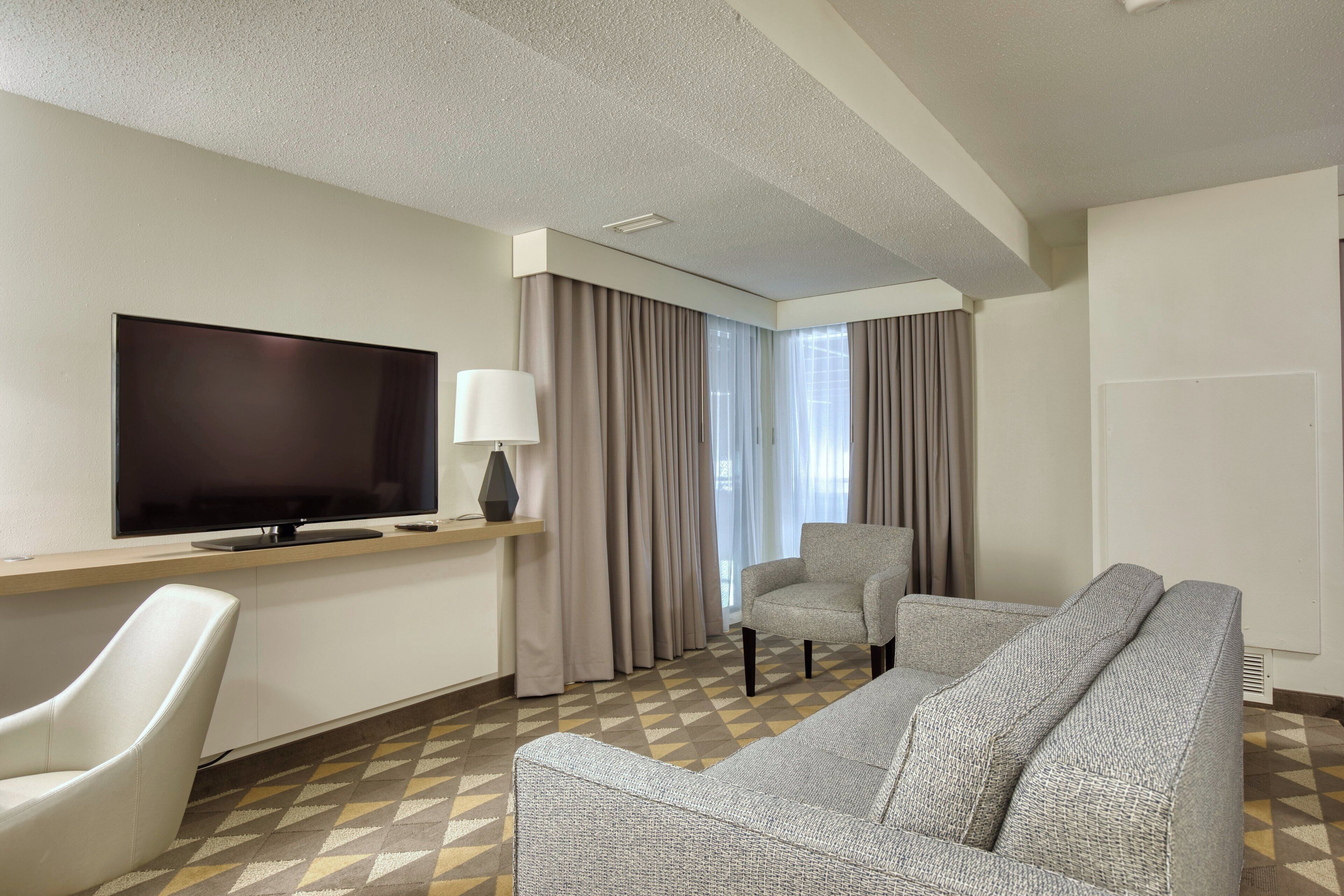 Habitación de hotel con televisión de pantalla plana, sofá, sillón, lámpara y ventana con cortinas.