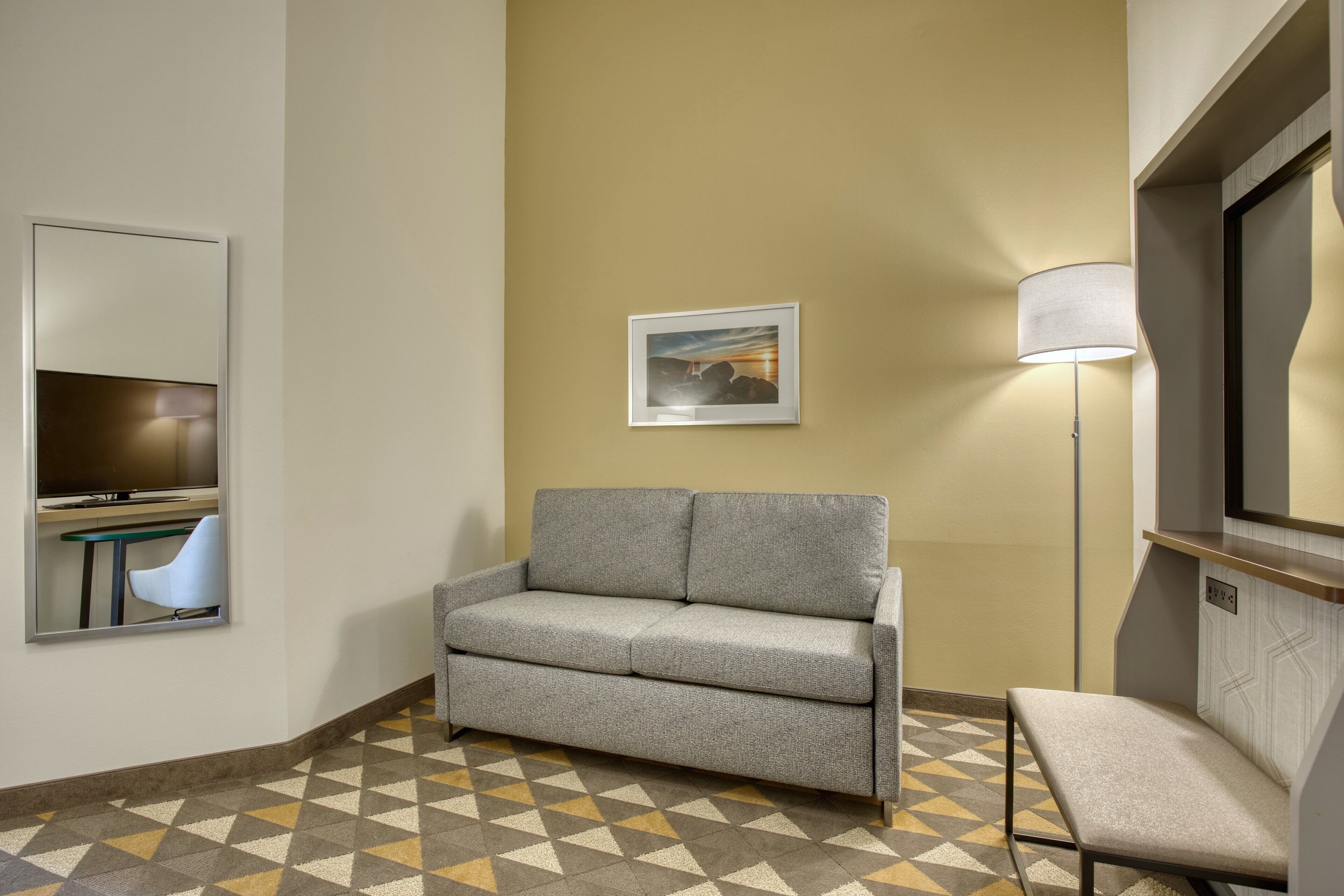 A hotel room with a sofa, a chair, a lamp, a framed picture on the wall, and a patterned floor.
