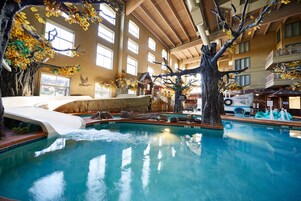 An indoor swimming pool with a waterfall feature, surrounded by artificial trees and a thatched-roof structure.