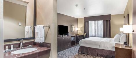 Habitación de hotel con una cama grande, un escritorio con televisor de pantalla plana, baño con lavamanos y espejo, y alfombra estampada.