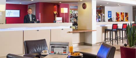 Eine Hottelobby mit einer Rezeption, einer Sitzecke mit Ledersesseln, einem Laptop auf einem Tisch und einer Bar im Hintergrund.