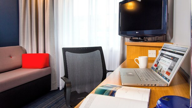 Ein Hotelzimmer mit einer Couch, einem Schreibtisch mit Laptop, einem Sessel, einem Fernseher und einem Fenster mit Vorhängen.