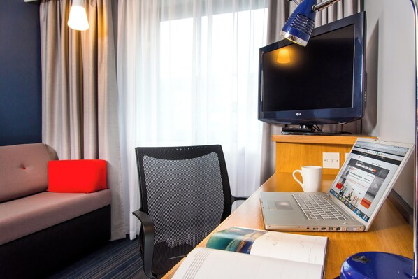 A hotel room with a sofa, a desk with a laptop, a chair, a TV, and a window with curtains. - Holiday Inn Express East Midlands Airport by IHG (Derby)