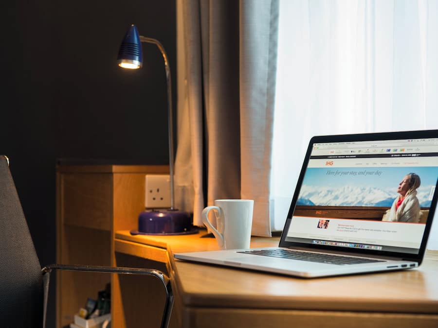 En bärbar dator står på ett skrivbord i trä, bredvid den finns en vit mugg. Till vänster står en lampa, och i bakgrunden syns ett fönster med gardiner.