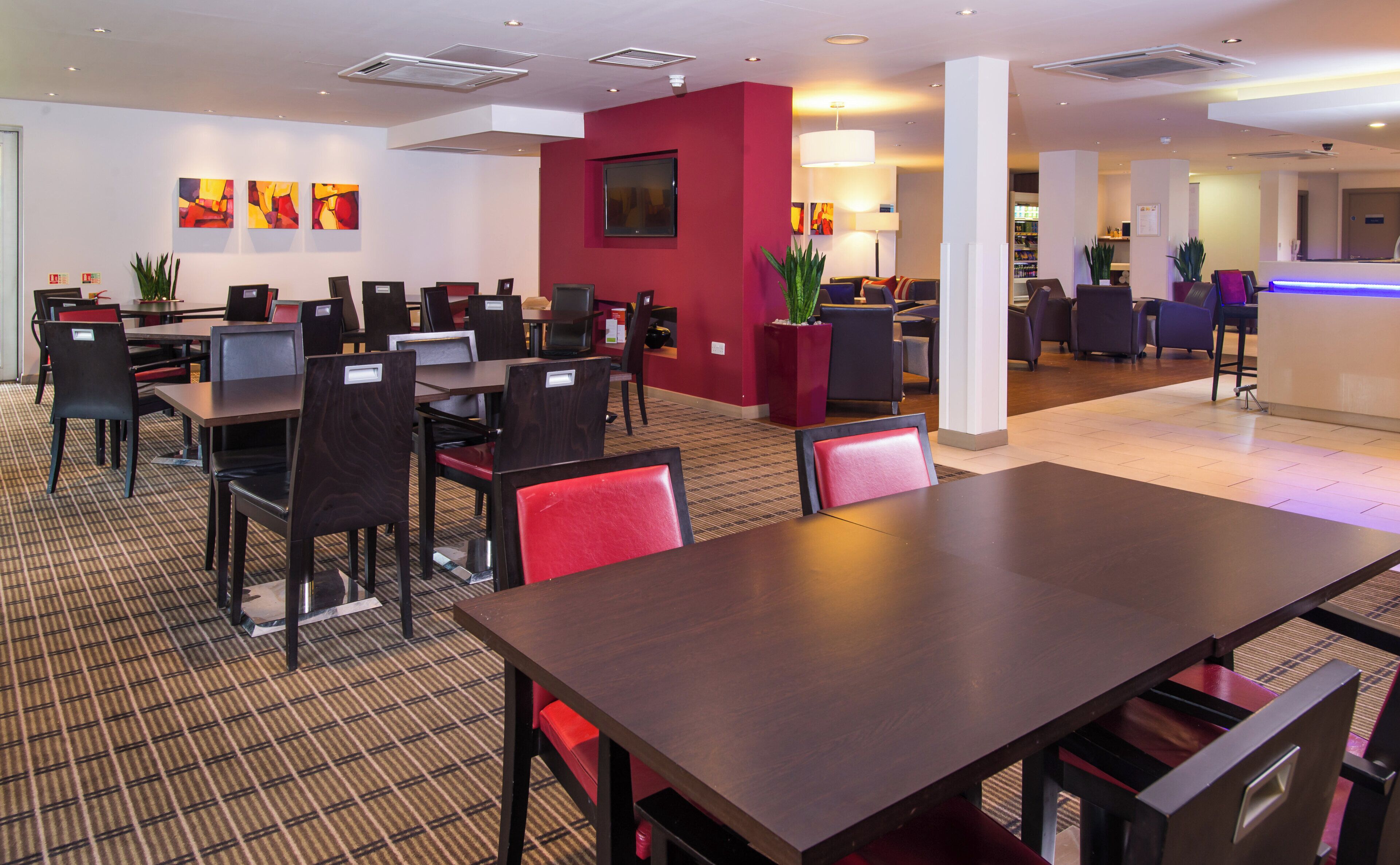 Un comedor de hotel con varias mesas y sillas, una pared de acento roja y una pantalla de televisión.