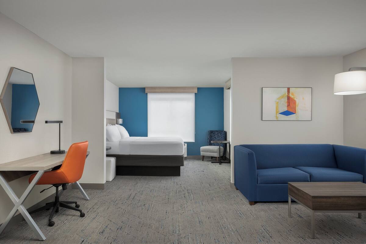 Une chambre d’hôtel avec un lit, un bureau avec une chaise orange, un canapé bleu, une petite table et un tableau au mur.