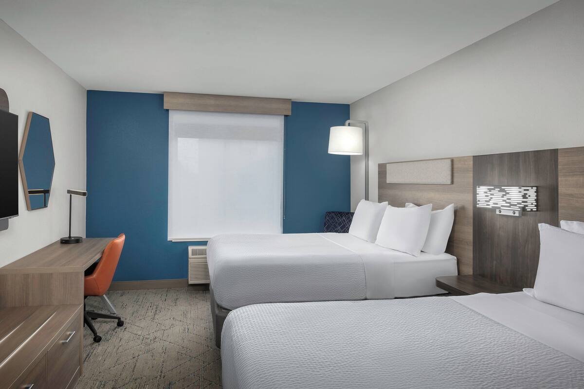 Une chambre d’hôtel avec deux lits, un bureau, une chaise, une télévision et une fenêtre avec des stores.