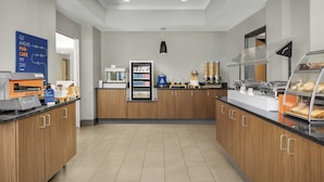 Une cuisine moderne dotée d’un espace où les clients peuvent se servir eux-mêmes pour le petit-déjeuner, comprenant une machine à café, un réfrigérateur et un présentoir avec des pâtisseries.