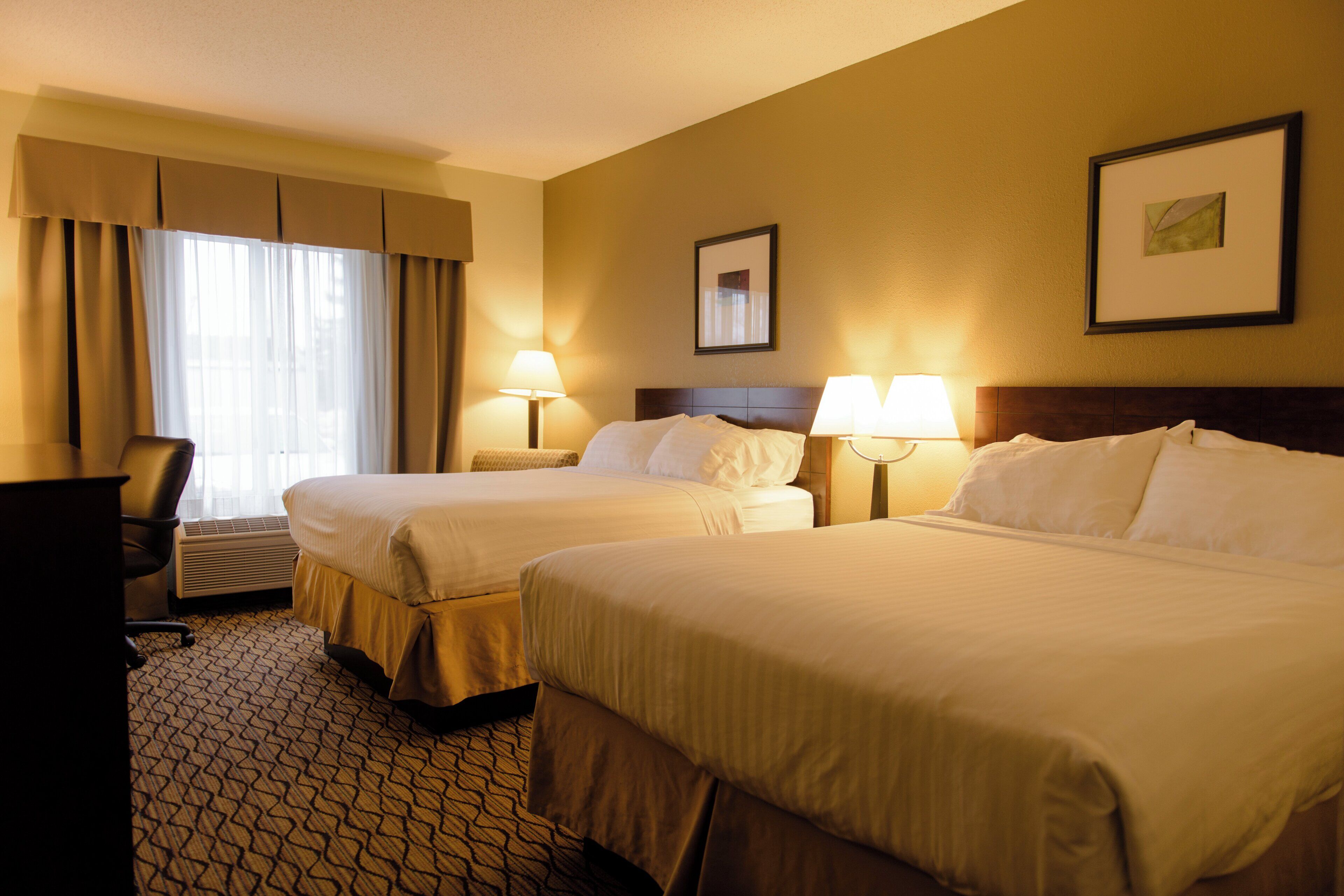 Ein Hotelzimmer mit zwei Betten, einem Schreibtisch und einem Sessel. Auf beiden Seiten der Betten stehen jeweils Lampen.