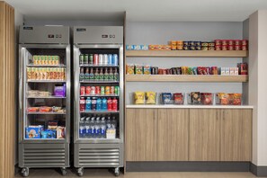 Duas geladeiras espaçosas repletas de bebidas e lanches, e prateleiras de madeira acima com diversos alimentos embalados.