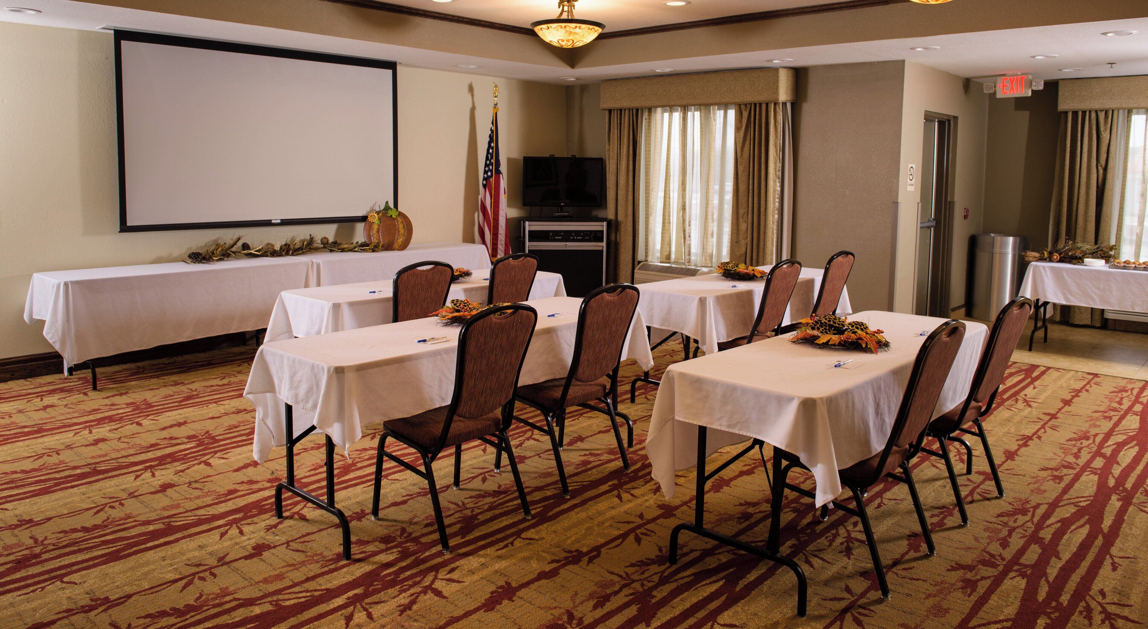A conference room with tables, chairs, a projector screen, an American flag, and a television.