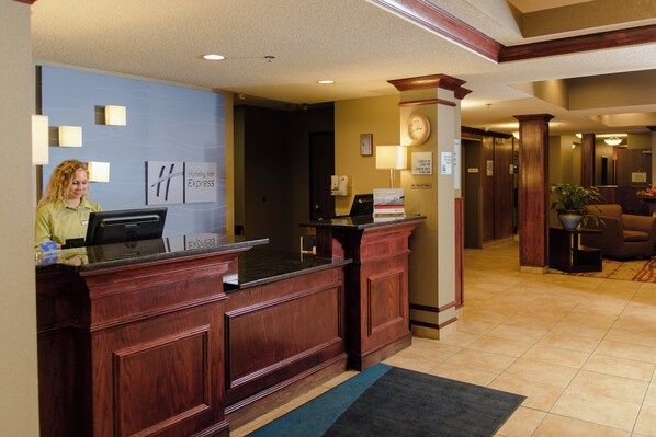 A hotel reception desk with a receptionist, a clock, and a sign indicating check-in times.