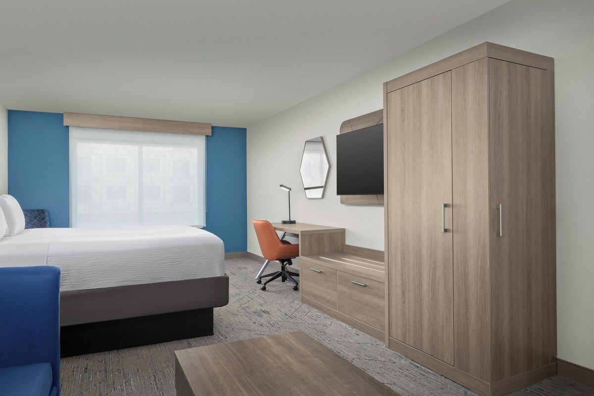Quarto de hotel com cama, escrivaninha, cadeira, armário e televisão fixada na parede.