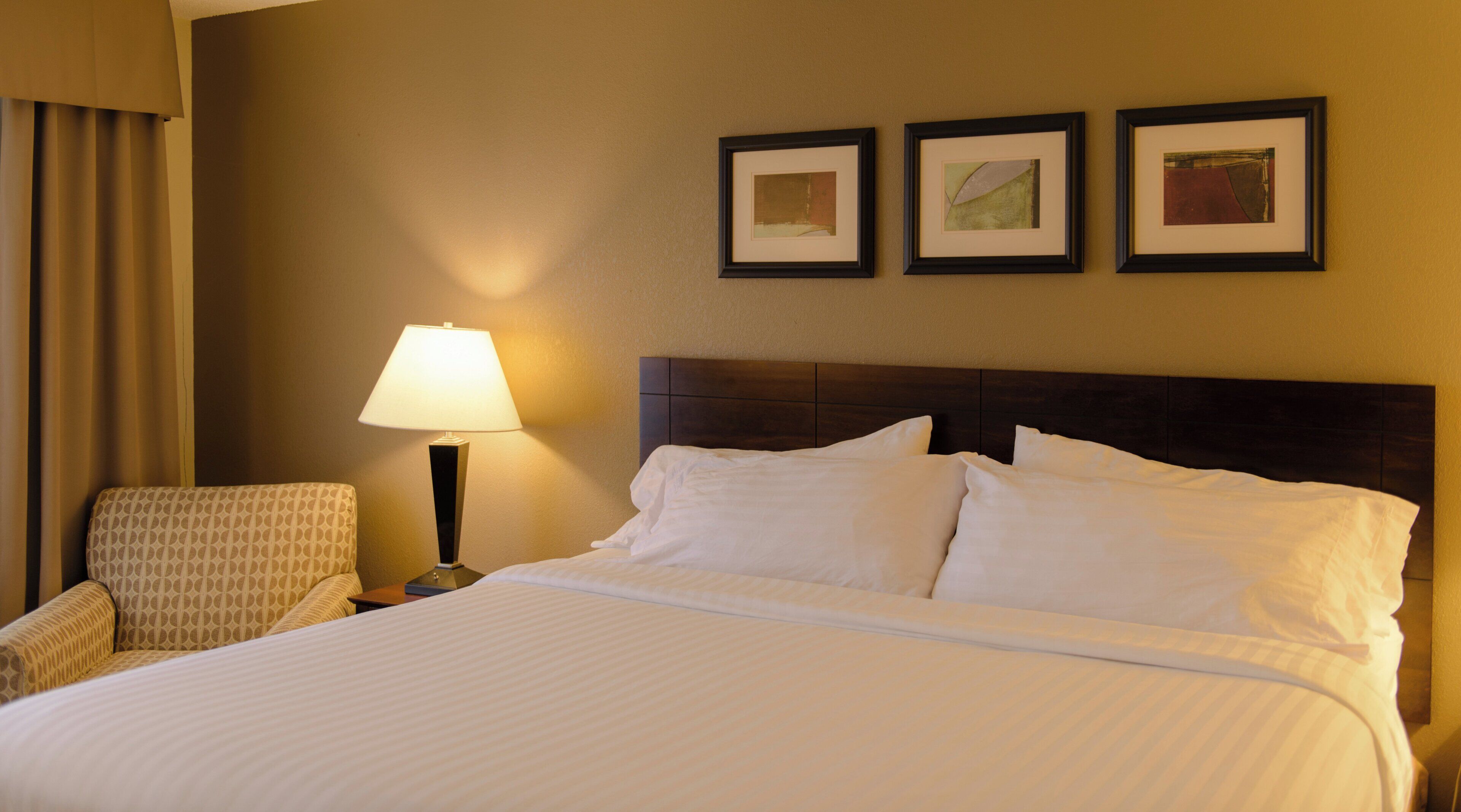 Habitación de hotel con una cama grande, una silla, una mesita de noche con lámpara y tres cuadros en la pared.