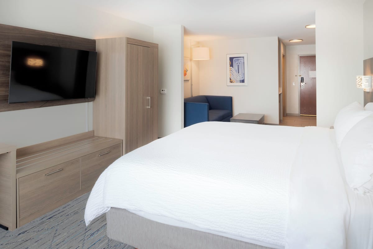 A hotel room with a large bed, a TV mounted on the wall, a desk with drawers, a chair, and a small table.