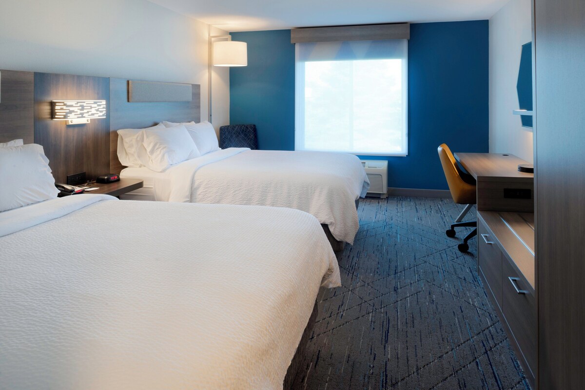 A hotel room with two beds, a desk, a chair, a lamp, and a window.
