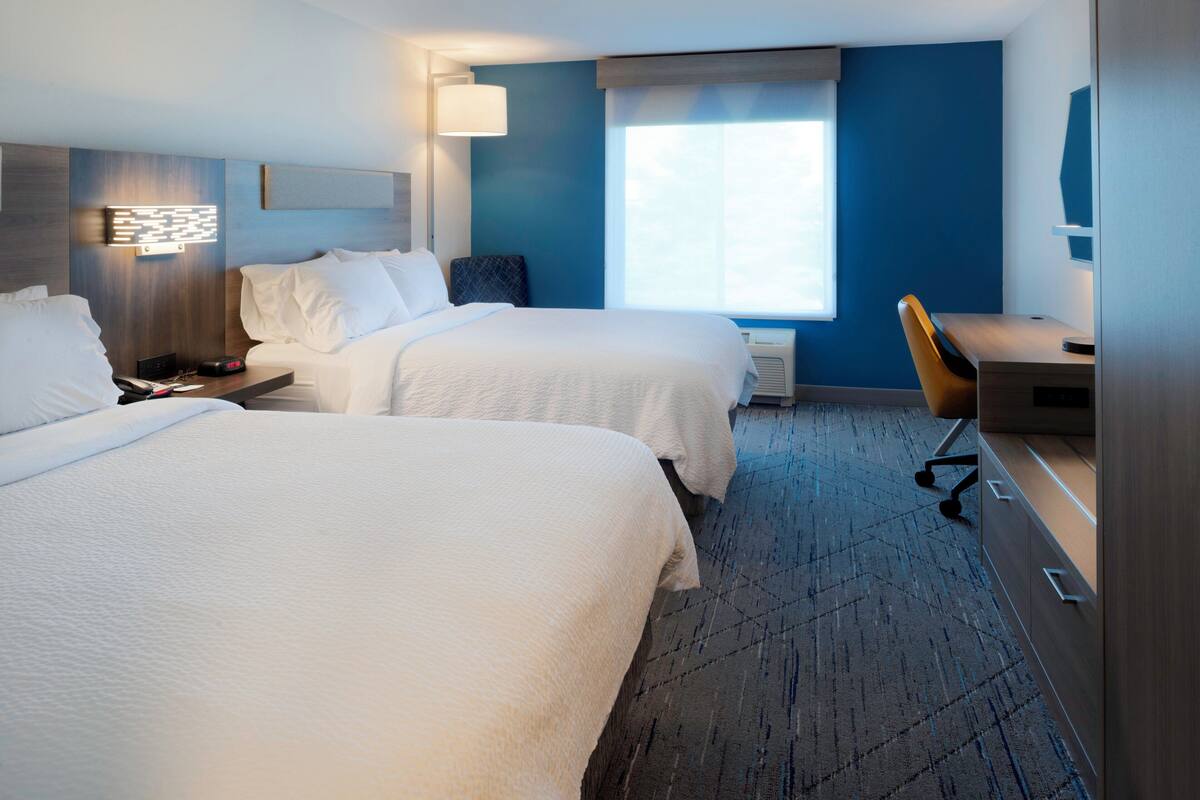 A hotel room with two beds, a desk, a chair, a lamp, and a window.