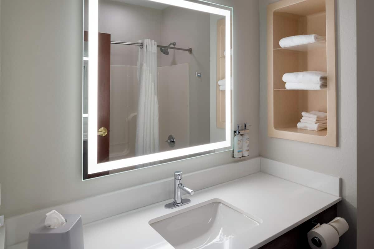 A bathroom with a large mirror, a white sink, a towel rack, and a shelf with folded towels.