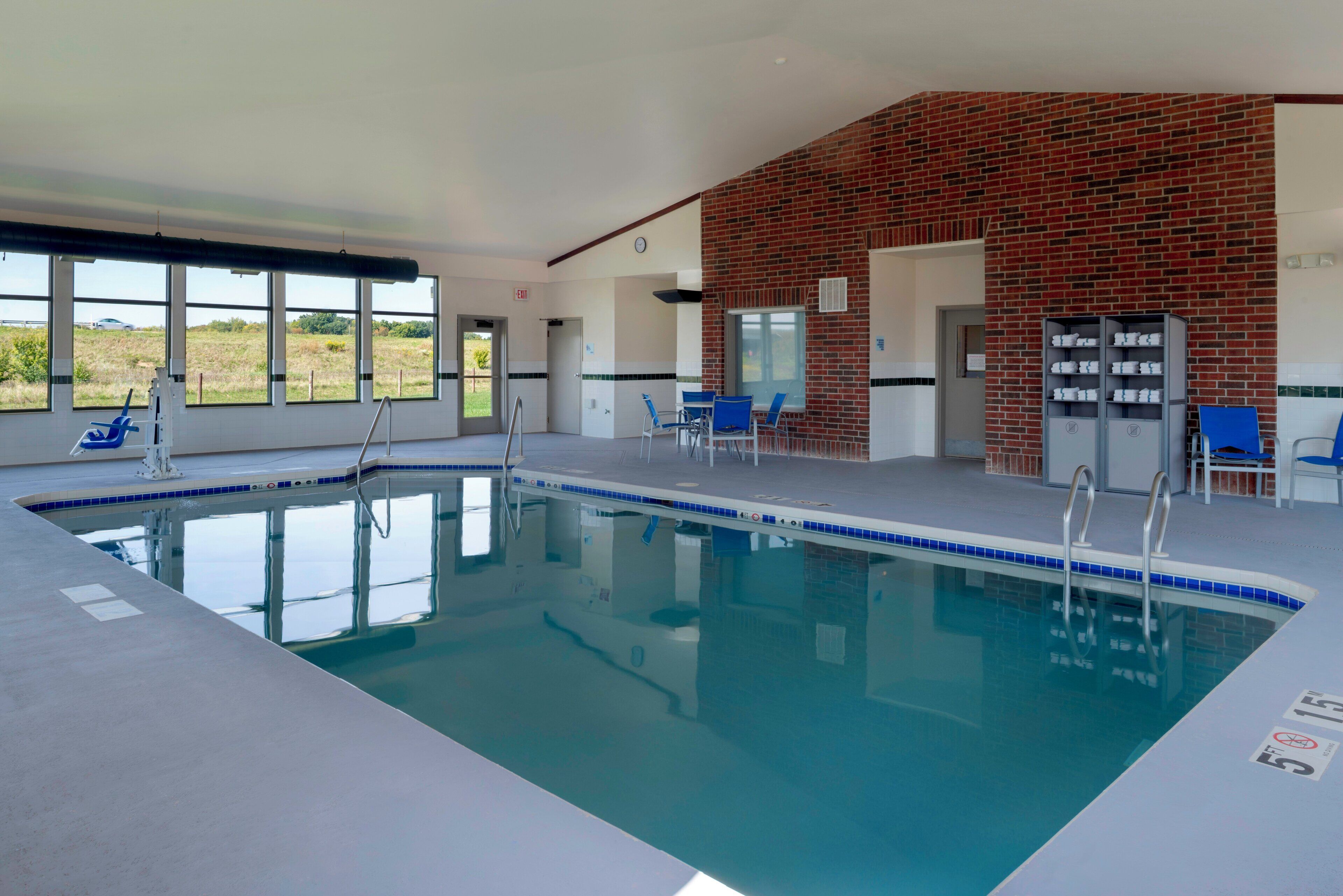 Una piscina interior con agua azul cristalina, rodeada de sillas azules y una pared de ladrillo.