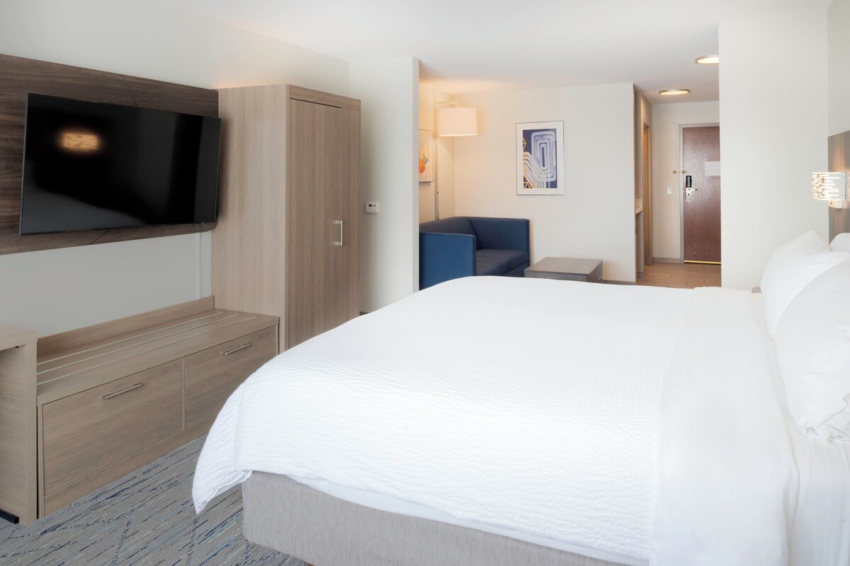 A hotel room with a large bed, a TV mounted on the wall, a desk with drawers, a chair, and a small table.