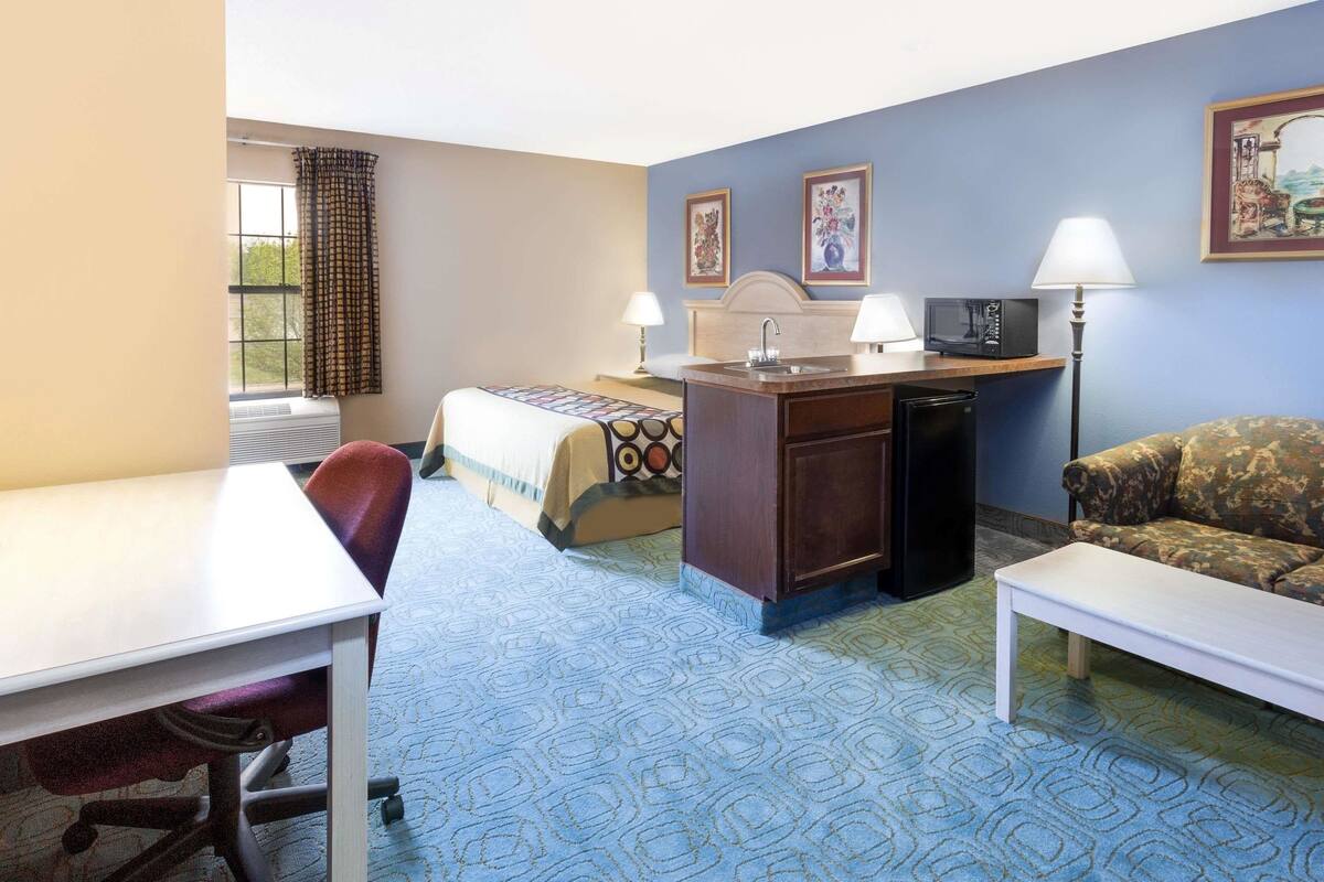 A hotel room with a bed, desk, chair, sofa, and a small kitchenette.