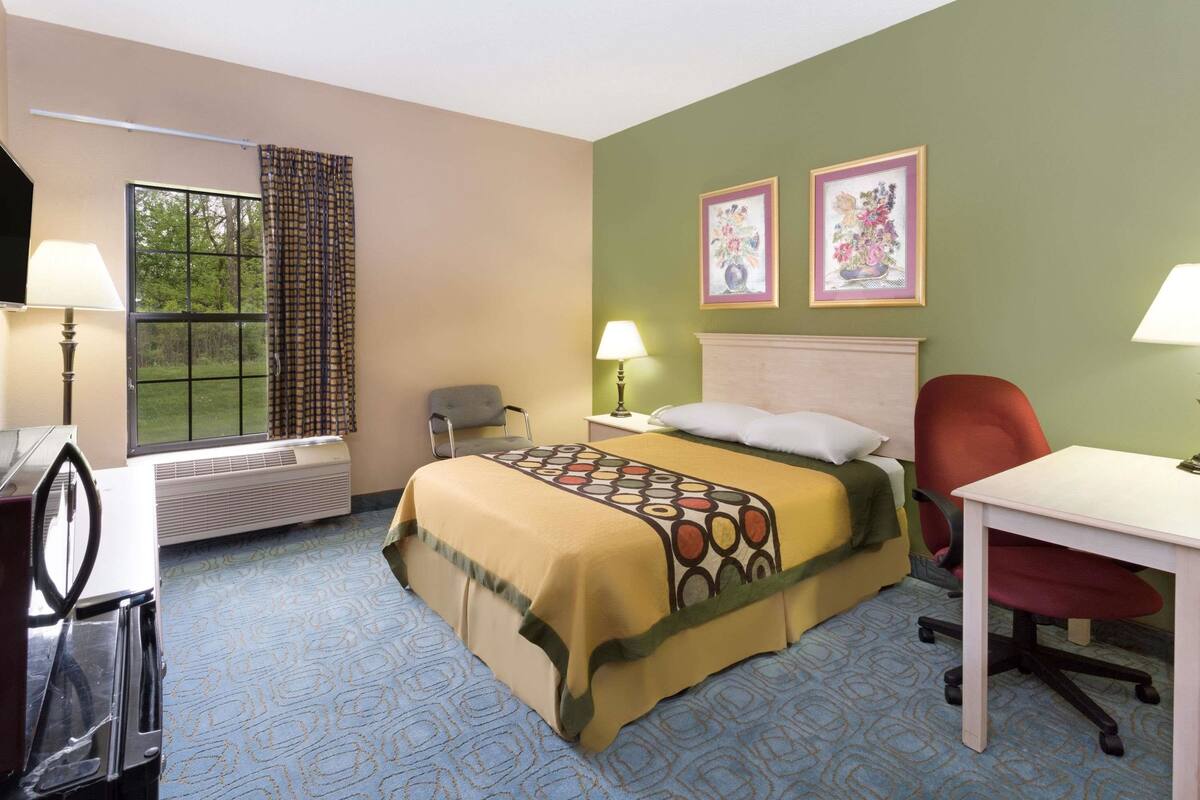 A hotel room with a bed, a desk, a chair, a window with curtains, and two framed pictures on the wall.