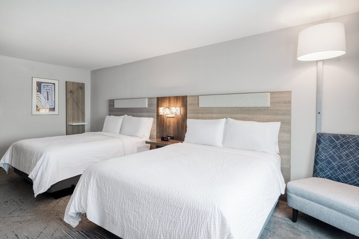 A hotel room with two beds, a nightstand, a lamp, and a wall-mounted artwork.
