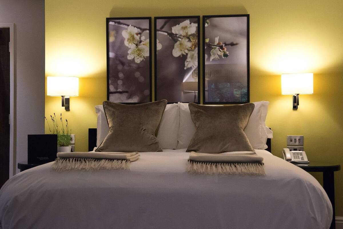 Habitación de hotel con una cama grande, dos lámparas fijadas a la pared y tres cuadros con motivos florales sobre la cama.