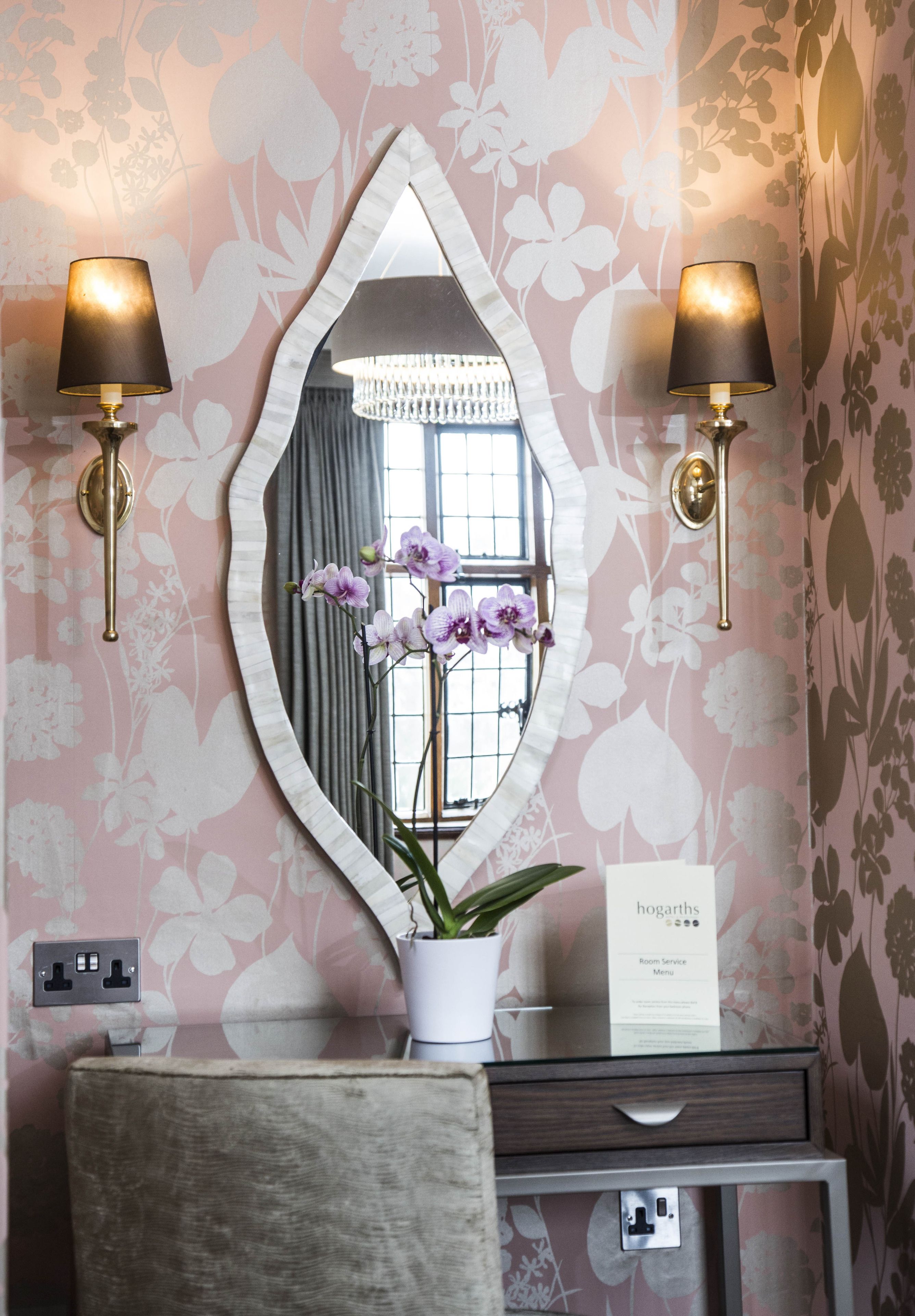 A room with a vanity, two wall-mounted lamps, a mirror, a potted orchid, and a sign indicating room service availability.
