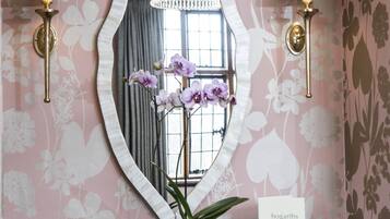 A room with a vanity, two wall-mounted lamps, a mirror, a potted orchid, and a sign indicating room service availability.