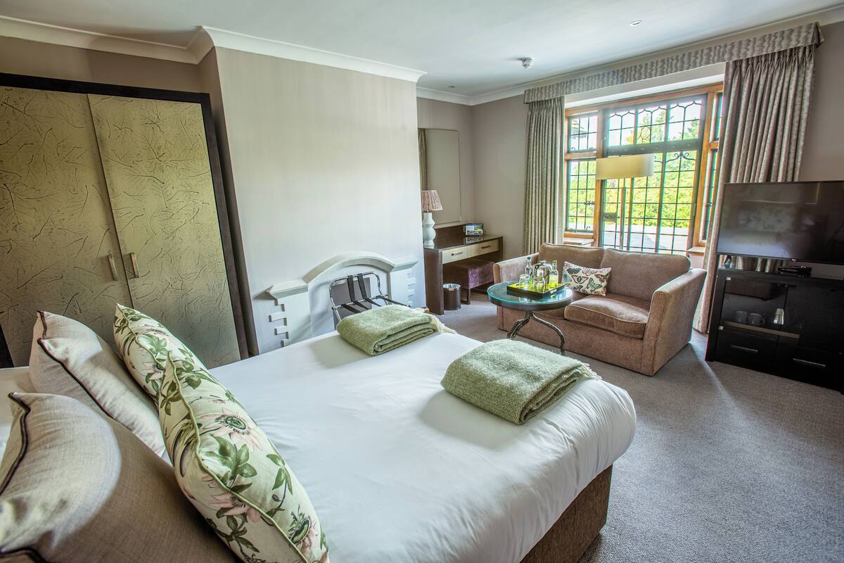 Habitación de hotel con una cama grande, una zona de estar con una silla y una mesita, televisión y ventana con cortinas.