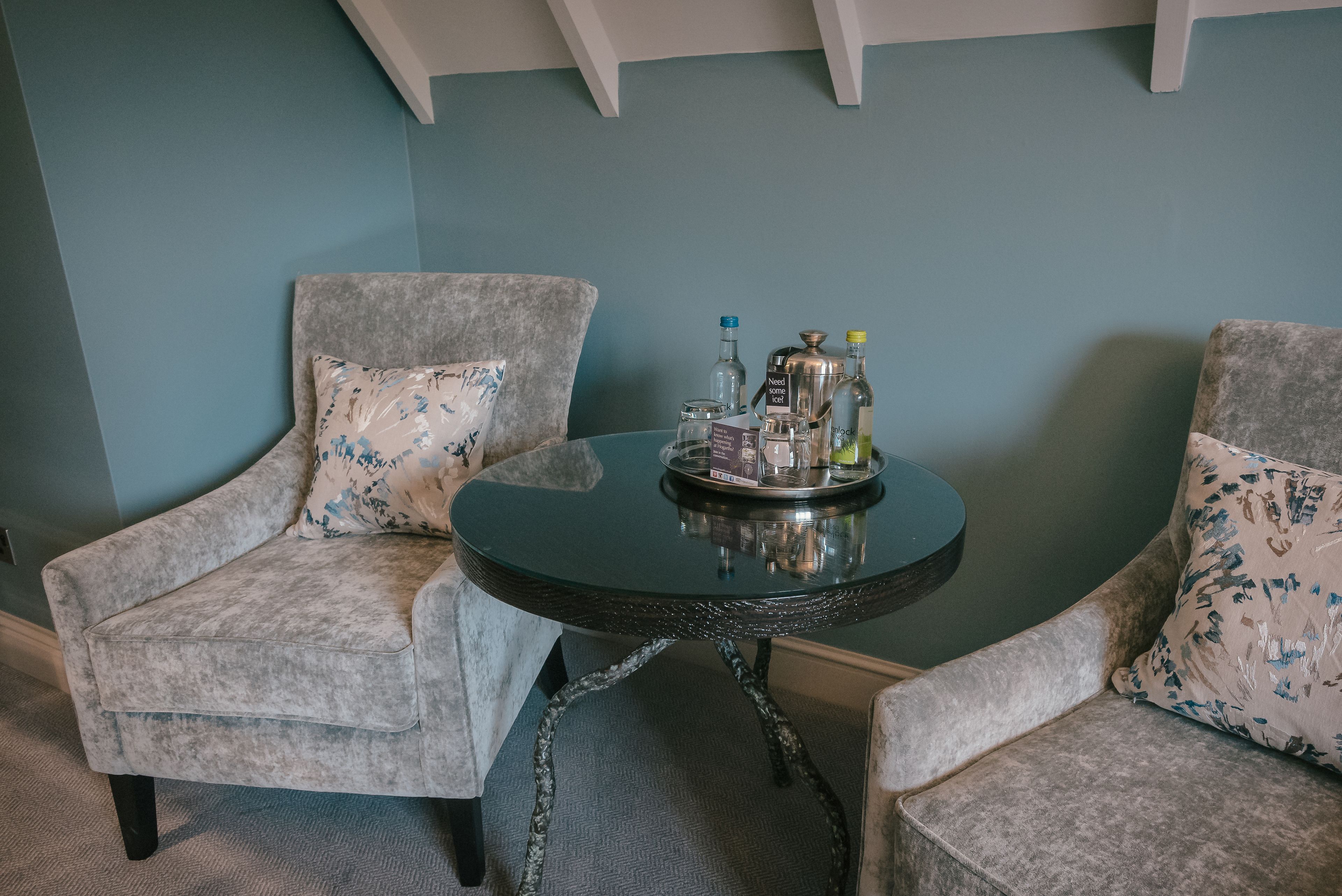 Et værelse med to grå polstrede stole med blomstermønstre, et rundt bord med metalstel og en bakke med forskellige flasker og en kaffekande.