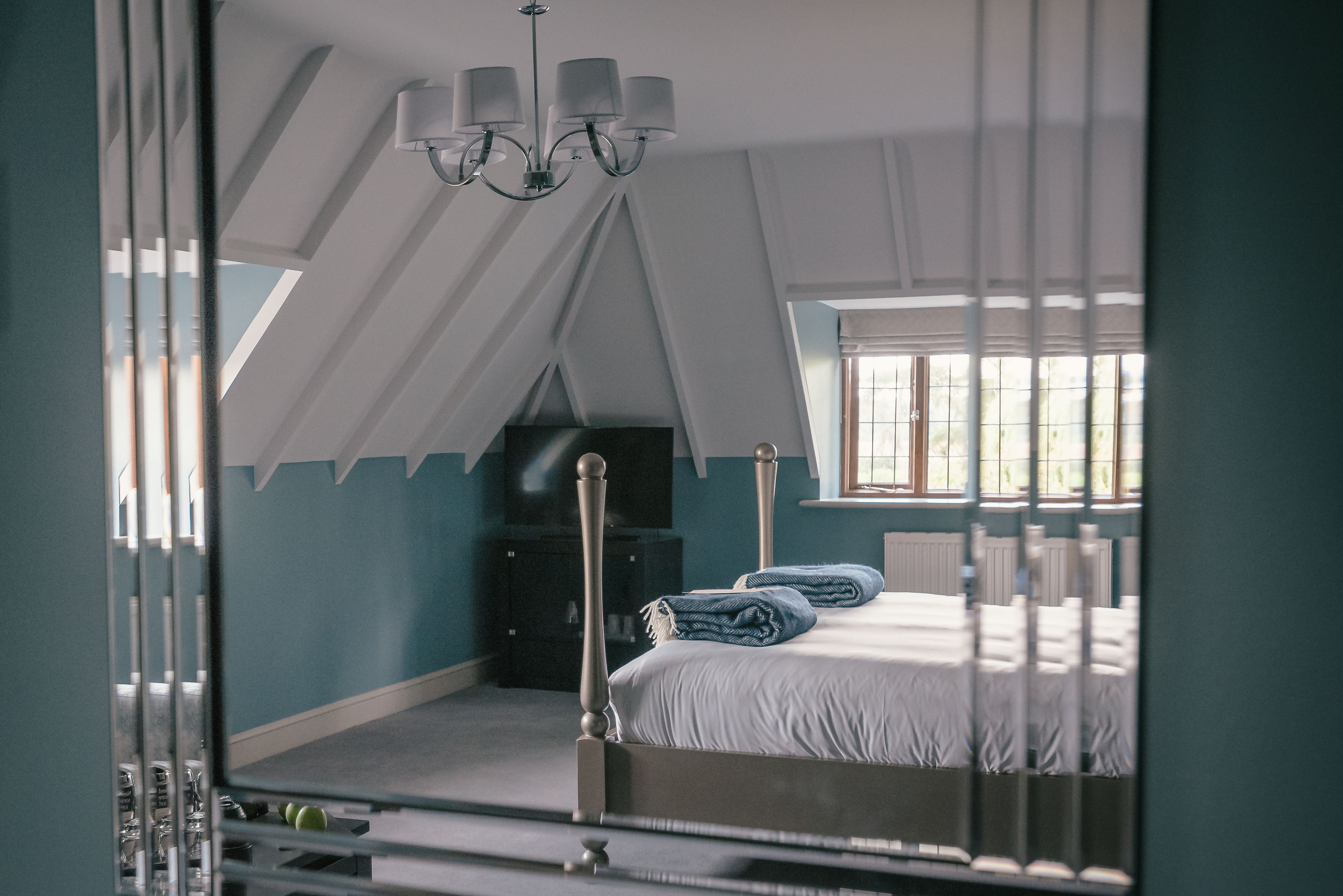 A bedroom with a sloped ceiling, a bed with white bedding, a chandelier, and a window with blinds.