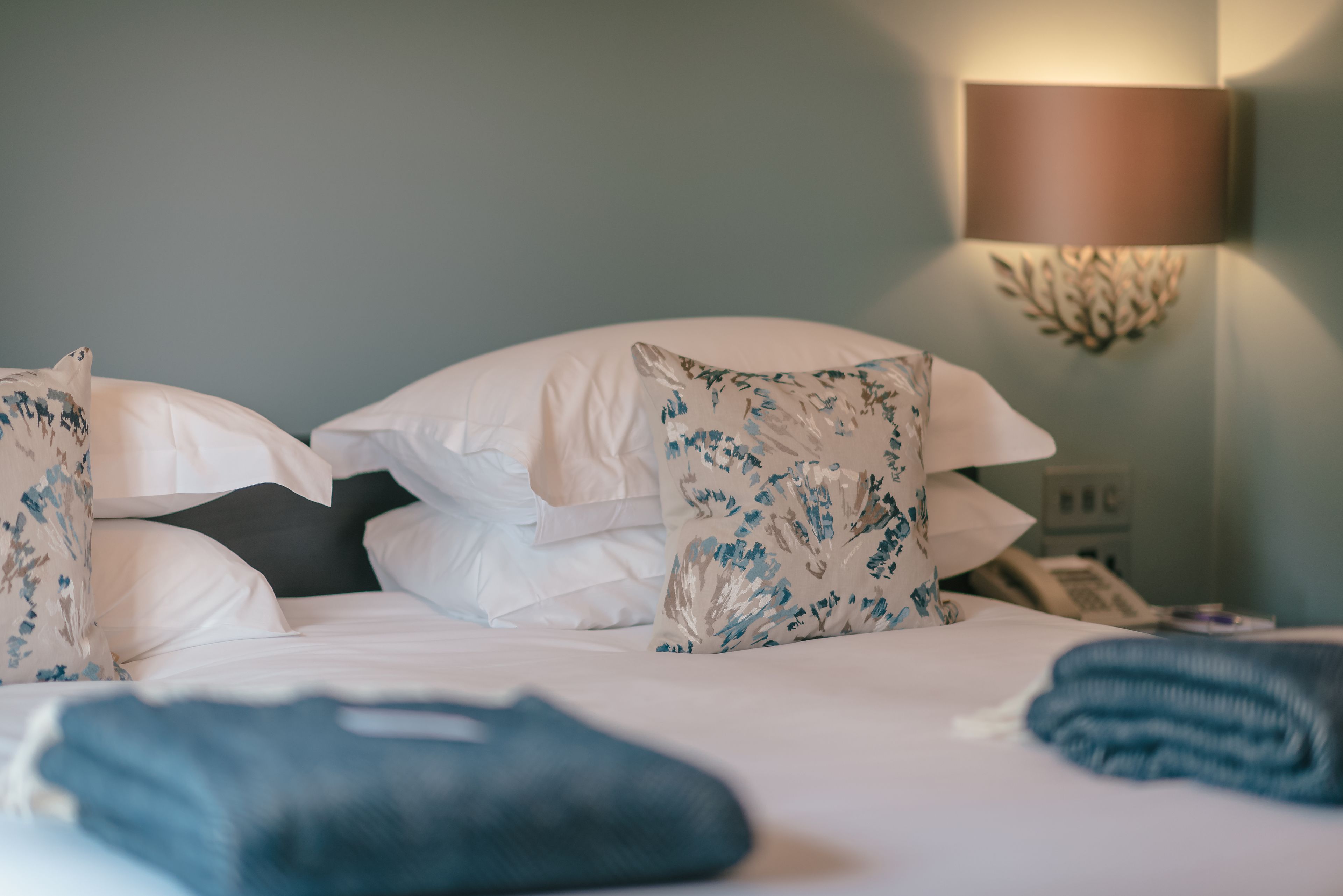A neatly made bed with white linens and decorative pillows, a bedside lamp, and a phone on the nightstand.