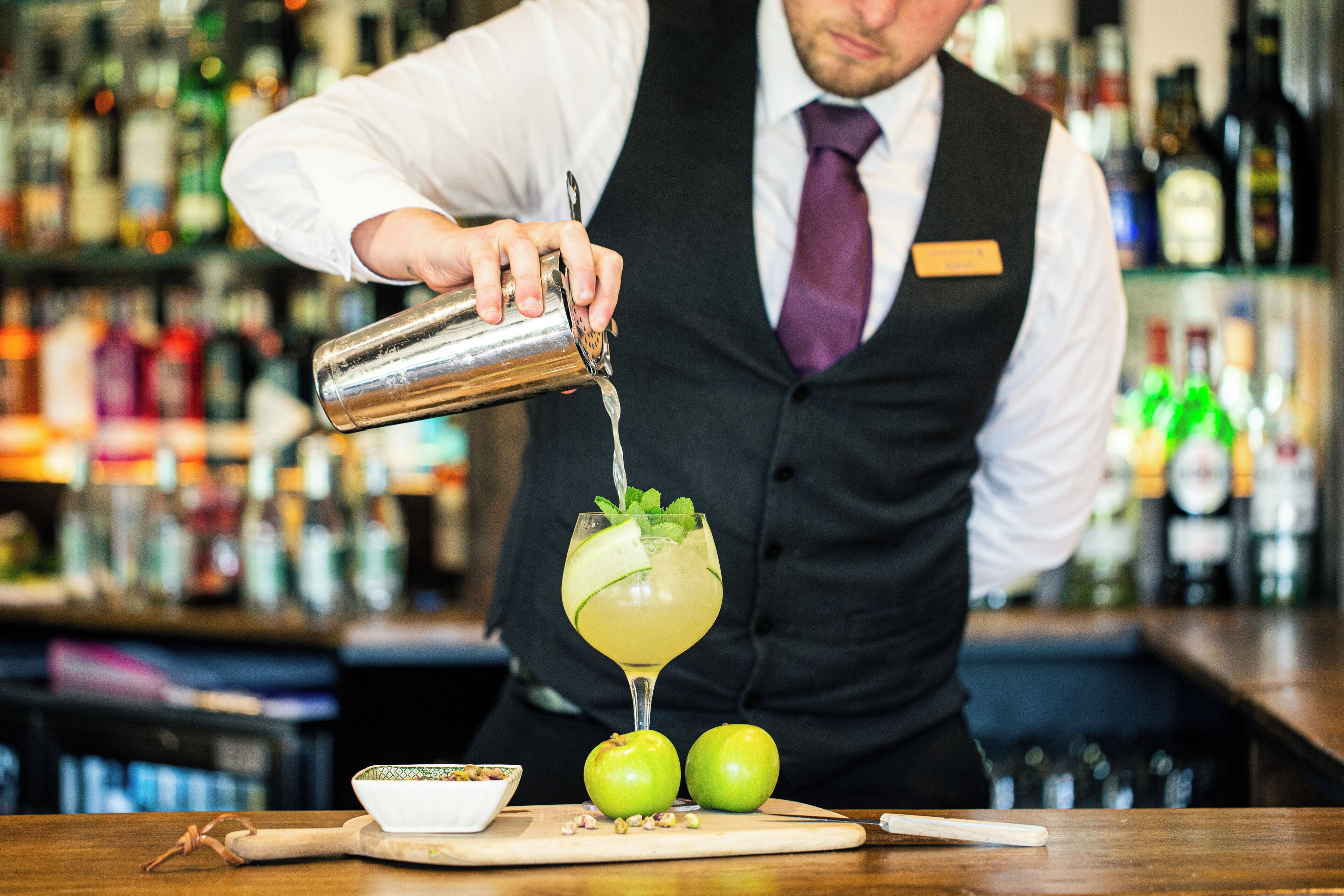 Een bartender schenkt een drankje in een glas, versierd met een partje citroen en munt. Op de bar liggen twee groene appels.
