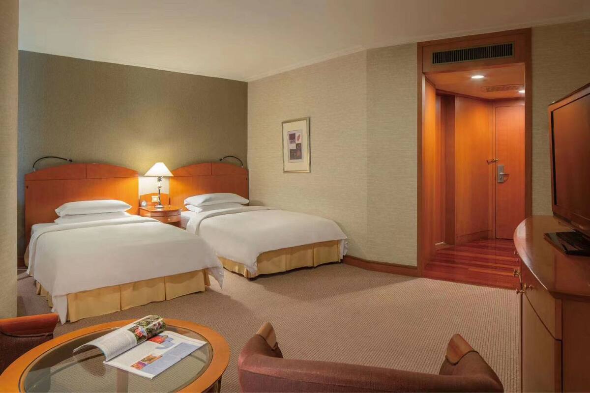 A hotel room with two beds, a desk with a lamp, a small table with a magazine, and a door leading to a hallway.