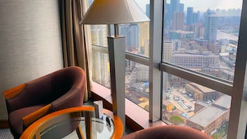 Une chambre d’hôtel dotée d’une grande fenêtre donnant sur la ville, d’une table ronde en verre, de deux fauteuils et d’un lampadaire.