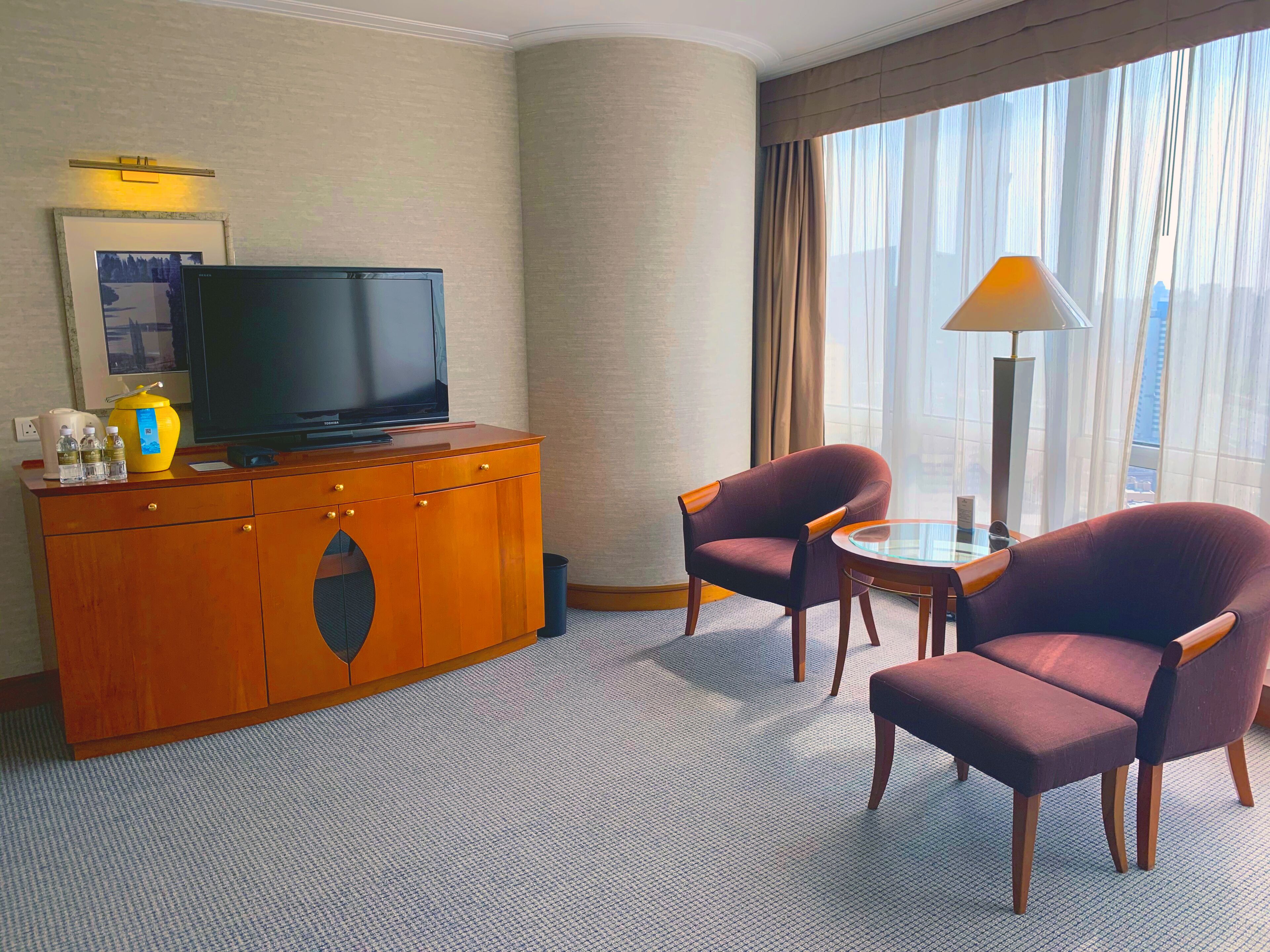Habitación de hotel con televisor de pantalla plana sobre un mueble de madera, dos sillones, una mesita y una lámpara.
