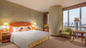 A hotel room with a large bed, two bedside tables with lamps, a green armchair, a small table with fruit, and a city view through large windows.