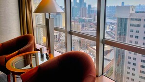 A room with a large window offering a city view, a chair, a table, and a floor lamp. - Hotel Nikko Dalian (Dalian)