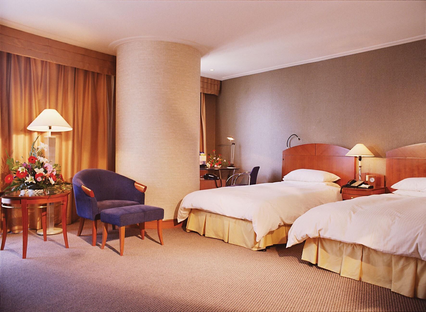 A hotel room with two beds, a desk, a chair, a lamp, and a flower arrangement.