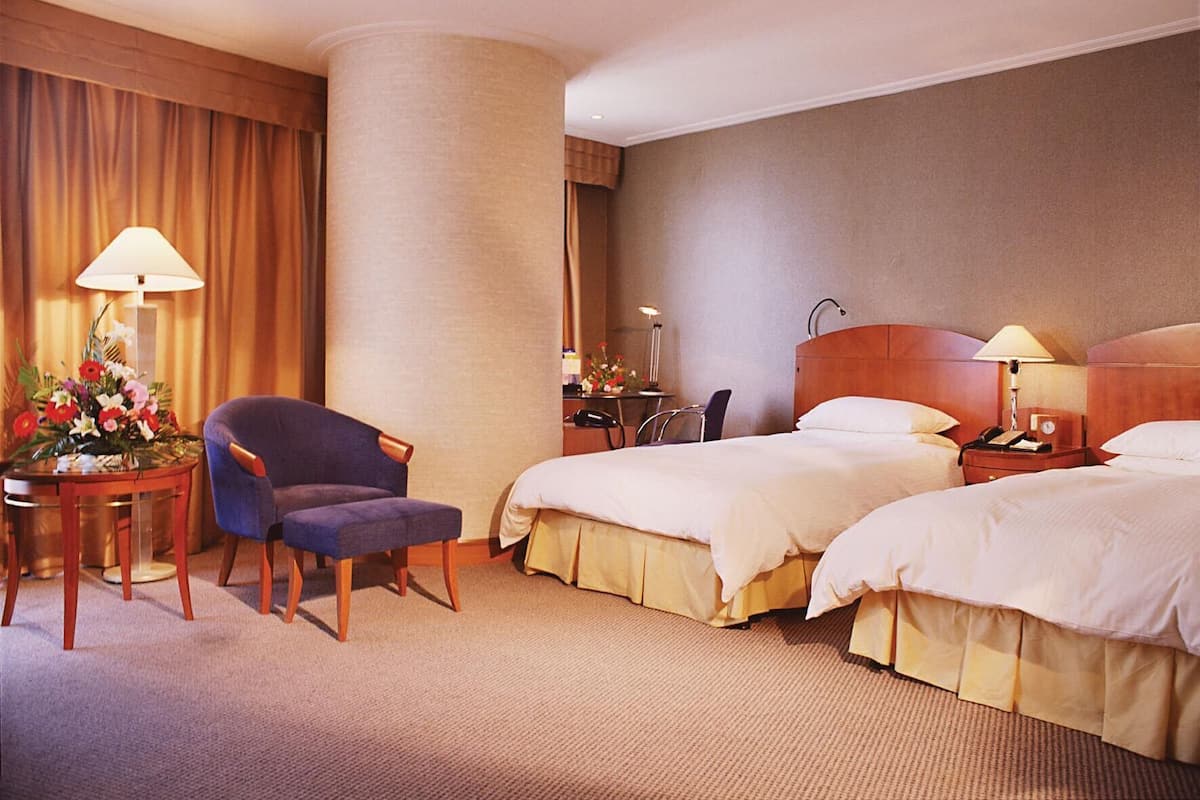 A hotel room with two beds, a desk, a chair, a lamp, and a flower arrangement.