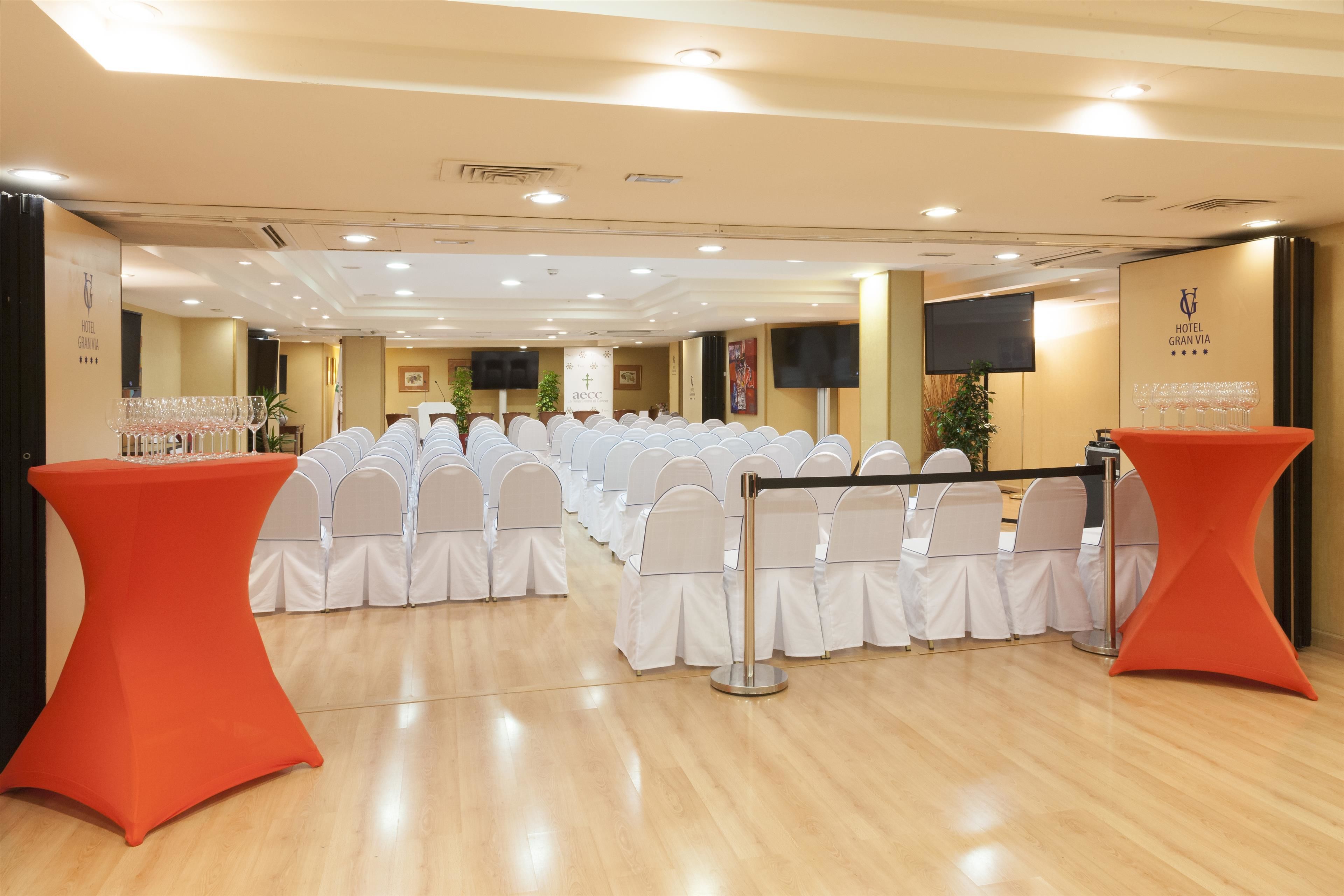 A hotel conference room with rows of white chairs, a red table, and a black rope barrier.