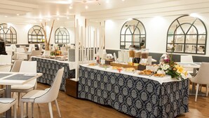 A dining area with tables and chairs, a buffet table with food and drinks, and large windows with arched tops.