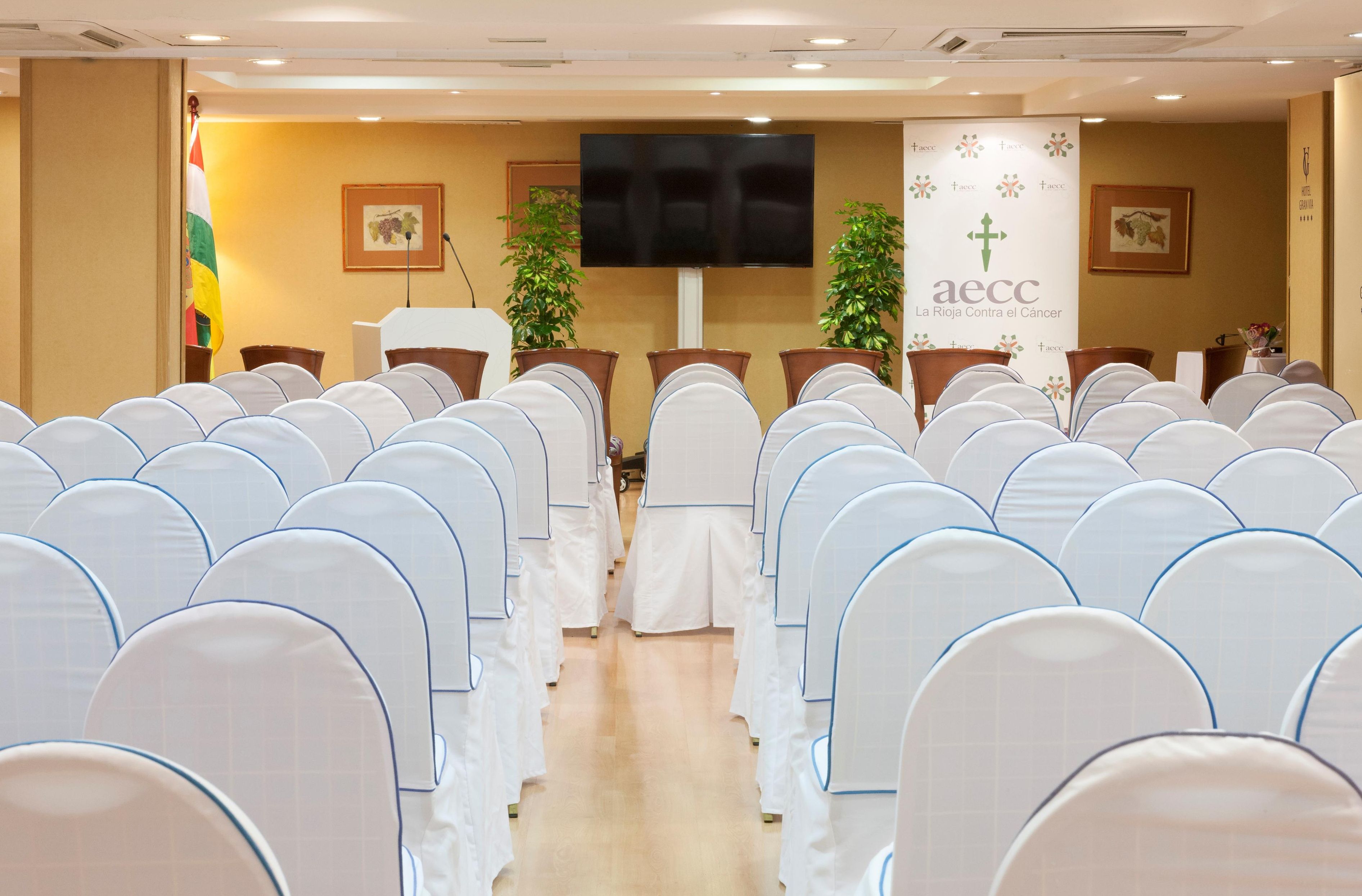 A conference room with rows of white chairs, a podium, a large screen, and banners.
