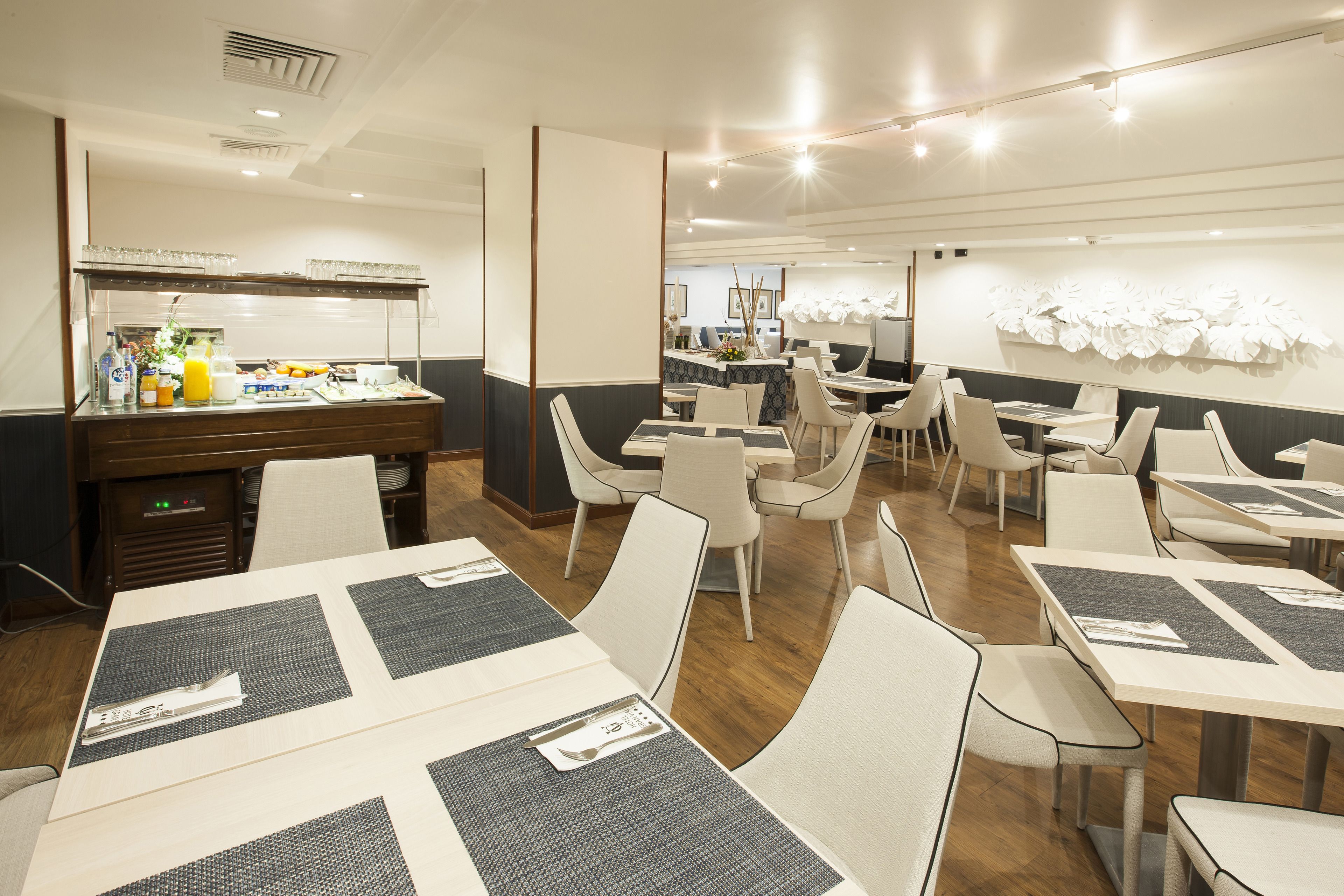 A modern dining area with tables and chairs, a buffet counter with food and beverages, and a decorative wall with white flowers.