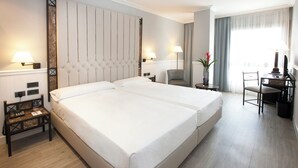 A hotel room with a large bed, two bedside tables, a desk with a chair, and a window with curtains.