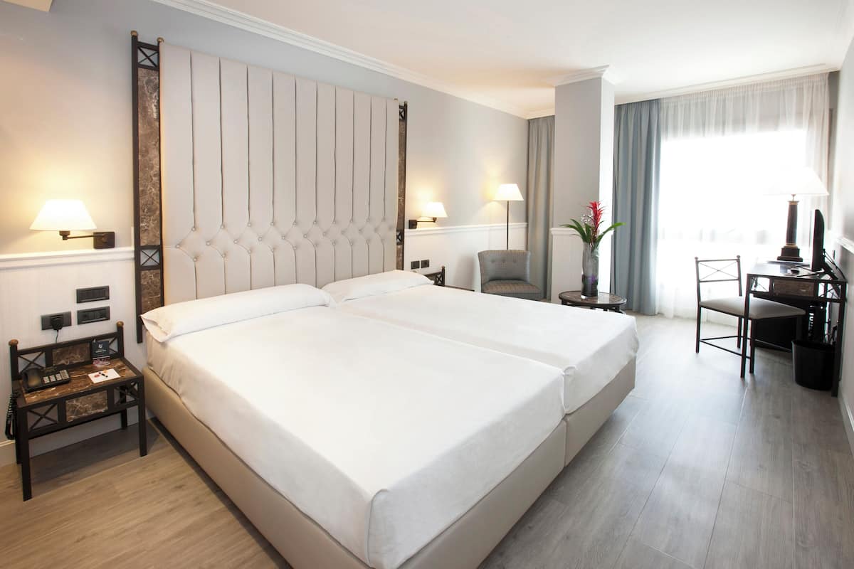 A hotel room with a large bed, two bedside tables, a desk with a chair, and a window with curtains.