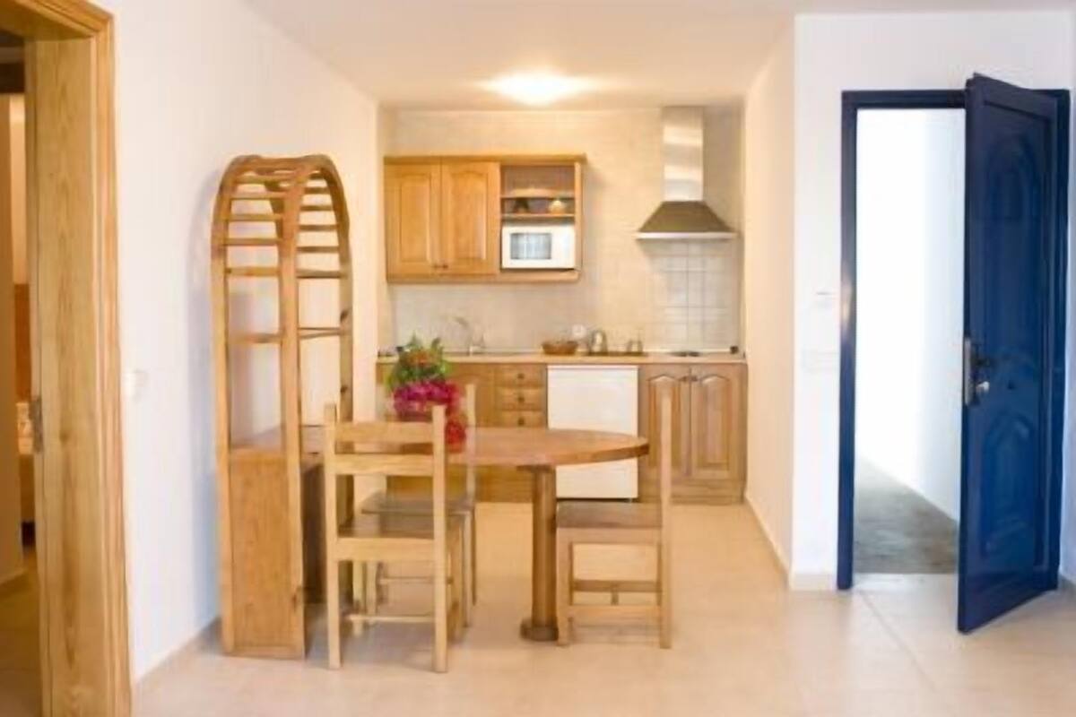 Cocina compacta con armarios de madera, microondas y un pequeño comedor con mesa y sillas.